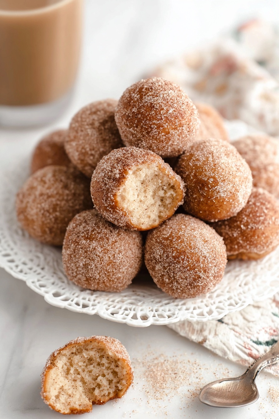 A pile of small round doughnut holes covered in cinnamon sugar sits in the center of a white plate with a floral lace edge design. The doughnut holes are golden brown with a fine layer of cinnamon sugar that looks slightly grainy. One doughnut hole, placed in the front on a white marbled surface, is split open, showing a soft, airy, light beige inside texture. A silver spoon with some cinnamon sugar sits nearby, and a glass of light brown drink is slightly out of focus in the background. photo taken with an iphone --ar 2:3 --v 7
