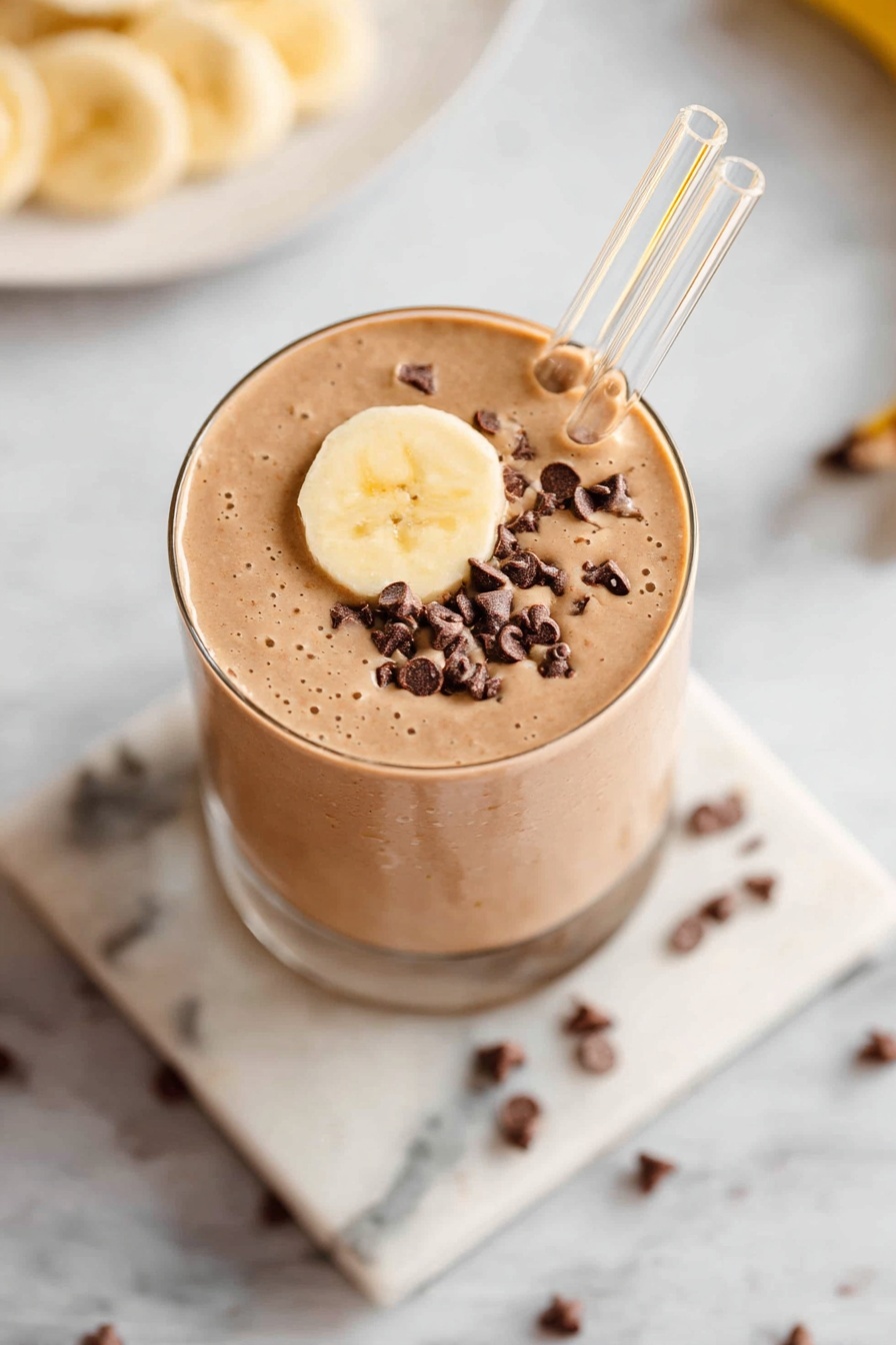 A glass filled with a smooth light brown smoothie topped with a round banana slice and small dark chocolate chips on one side of the surface. Two clear glass straws stick out from the top, angled slightly apart. The glass is placed on a square white marble coaster with grey veins, and the background is a white marble surface with scattered chocolate chips nearby. A glimpse of a white plate with more banana slices is partially visible at the top left corner. Photo taken with an iphone --ar 2:3 --v 7