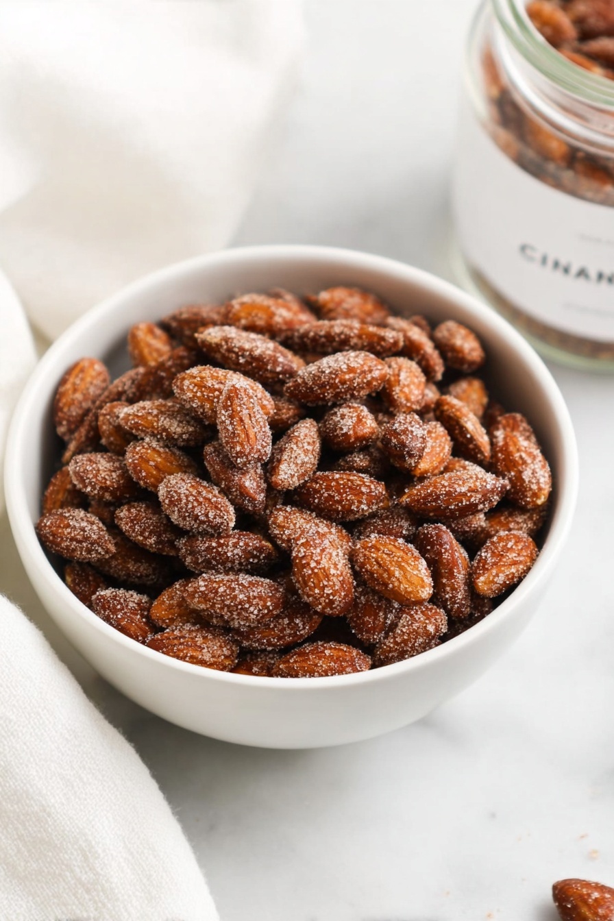 A white bowl filled with a single layer of roasted almonds covered in a coarse cinnamon sugar coating, giving them a rough texture and a light brown color. The bowl sits on a white marbled surface, surrounded by a soft white cloth and a clear jar labeled