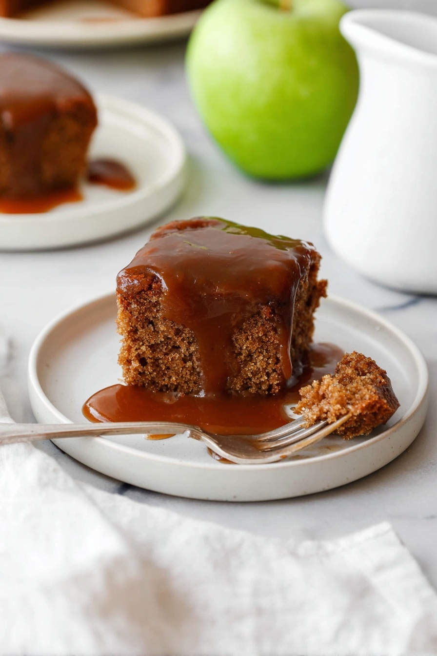 A piece of brown cake sits on a small white round plate on a white marbled surface. The cake has a rough texture and is topped with a thick, dark caramel sauce covering the top and partially dripping down the sides. In front of the cake on the plate, a small piece is cut and a silver fork is stuck into it, showing the moist inside of the cake with caramel sauce pooling underneath. In the background, a green apple and a white pitcher are slightly blurred, along with another plate with cake on the same white marbled surface. A white cloth is partly visible in the lower frame, adding softness to the scene. Photo taken with an iphone --ar 2:3 --v 7