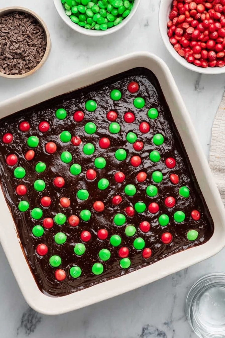 The image shows a close-up view of square dessert bars arranged in a grid. Each square has three layers: a dark brown bottom layer that looks like a dense brownie, a middle layer of white cream or frosting, and a top layer decorated with red and green candy-coated chocolates, small green and red round sprinkles, dark brown chocolate chunks, and tiny white snowflake-shaped sprinkles. The candies and sprinkles are spread unevenly on the white top layer, creating a colorful and festive look. The dessert squares rest on a dark wooden surface shown between the gaps. photo taken with an iphone --ar 2:3 --v 7